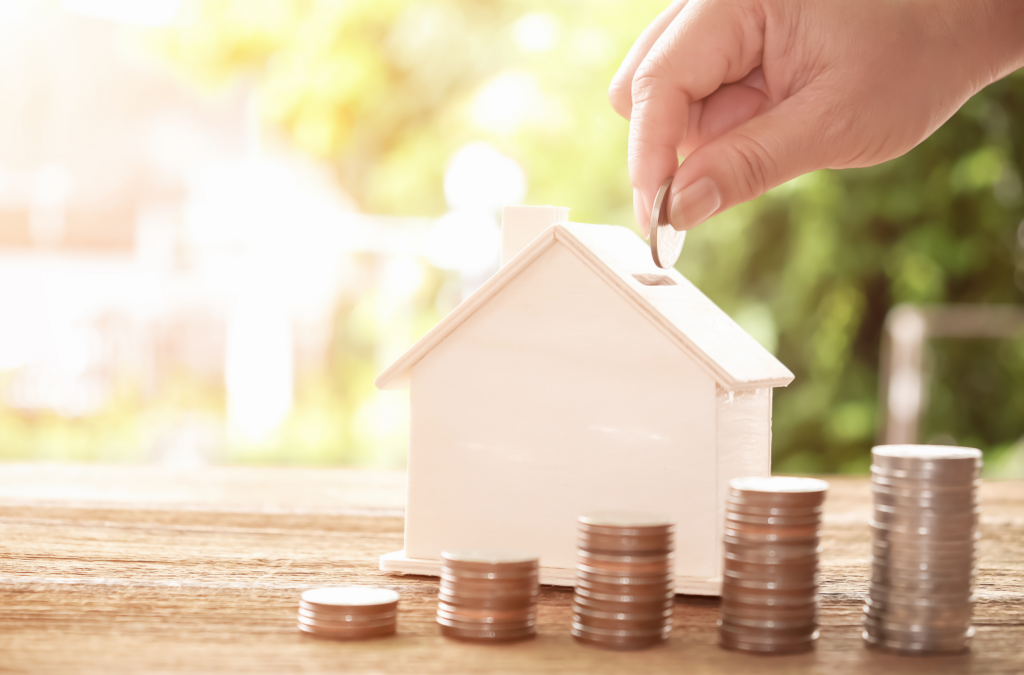 House money bank with coins in front and hand putting money in coin slot.